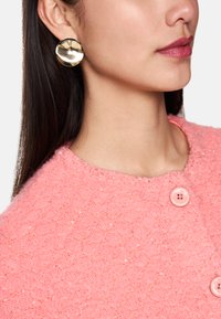 Rosa strukturierter Pullover mit einem Rautenmuster, ausgestattet mit zwei rosa Knöpfen am Hals. Goldene runde Ohrringe werden getragen.