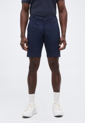 Modèle masculin portant un short bleu marine, des chaussettes blanches et des baskets blanches, debout devant un fond clair uni.