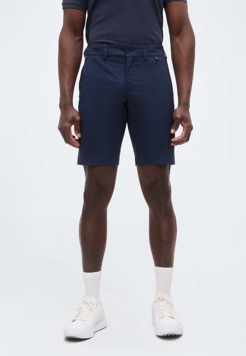 Άνδρας μοντέλο που φοράει σκούρα μπλε σορτς, λευκές κάλτσες και λευκά αθλητικά παπούτσια, στέκεται απέναντι σε ένα απλό φωτεινό φόντο.