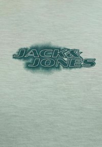 Πράσινο λογότυπο "JACK & JONES" με θολό φόντο τυπωμένο σε ανοιχτοπράσινο υφασμάτινο ύφασμα με υφή.