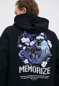 Mees, kes kannab musta kapuutsi, millel on lillakad koi kalad, kirsiõied, pagood ja tagaküljel tekst "Memorize".