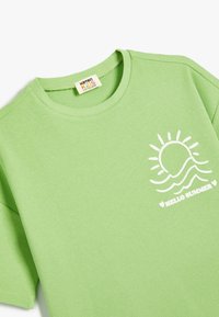 Vihreä ribbipaita, jossa on lyhyet hihat, ja etuosassa on valkoinen aurinko- ja aalto-kuvio sekä teksti "HELLO SUMMER".
