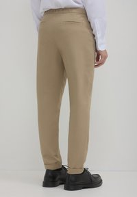 Beige Anzughose aus leichtem Stoff, mit einer flachen Gesäßtasche und Umschlägen am Saum, kombiniert mit schwarzen Schuhen.