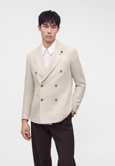 Jeune homme portant un blazer croisé beige avec des boutons dorés, une chemise blanche et un pantalon foncé sur un fond clair uni.
