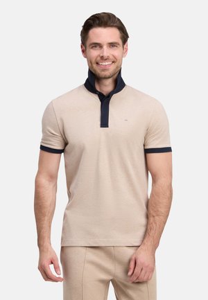 Homme souriant portant un polo beige à manches courtes avec un col et des bords de manches bleu marine foncé, debout devant un fond clair uni.