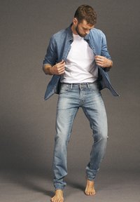 Lichtblauwe spijkerbroek gecombineerd met een licht denim overhemd over een wit T-shirt. Het model staat op blote voeten tegen een neutrale achtergrond.