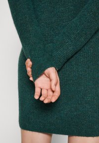 Maglione verde scuro a maglia con maniche lunghe, caratterizzato da una texture a coste e un design con polsini aperti. Le mani sono delicatamente unite dietro.