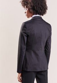 Schwarzer maßgeschneiderter Blazer mit einer taillierten Silhouette, strukturierten Schultern und glattem Stoff. Verfügt über einen Schlitzausschnitt hinten und einen hohen Kragen.