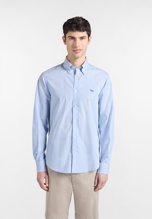 Junger Mann, der ein hellblau gestreiftes Hemd mit Knopfleiste und kleinem Hund-Logo sowie beige Shorts trägt, vor einem weißen Hintergrund stehend.