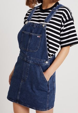 Frau trägt ein blaues Jeans-Overallkleid mit Front- und Seitentaschen über einem schwarz-weiß gestreiften Kurzarmshirt.