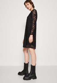 Black lace dress with long sleeves, floral pattern, and a loose fit. Paired with chunky black ankle boots for contrast.