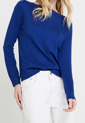 Kvinde iført en tætsiddende langærmet trøje i kongeblå, let stoppet ned i højtaljede hvide jeans med forlommer.