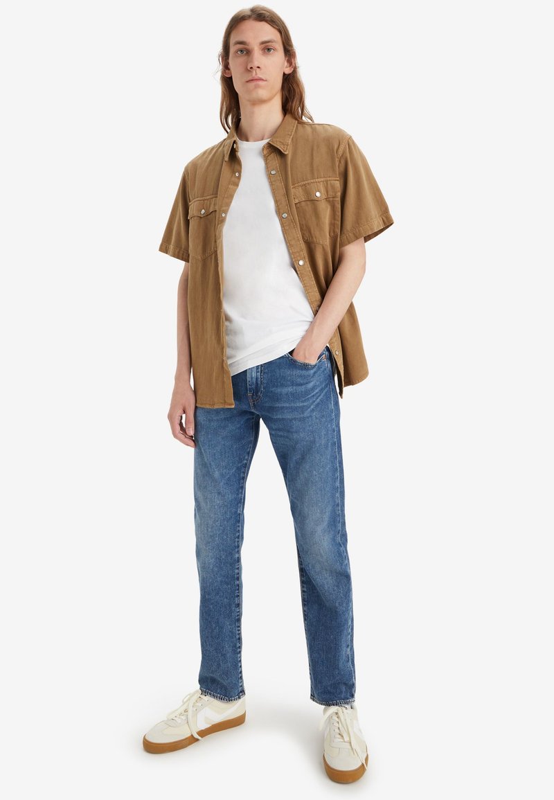 Braunes kurzärmliges Hemd über einem weißen T-Shirt, kombiniert mit blauen Jeans und weißen Sneakers. Das Hemd hat einen Knopfverschluss.
