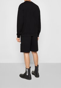 Felpa nera con vestibilità oversize, abbinata a pantaloni corti neri. Le calzature includono stivali in pelle nera con il marchio "Moschino" in bianco.