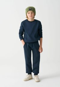 Tamsiai mėlynas džemperis ir suderinti kelnės iš minkštos medžiagos, derinami su žaliu kepuraite. Šviesios spalvos sportbačiai užbaigia aprangą.