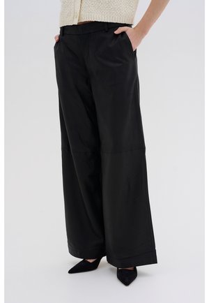 Persoon die zwarte, hoogtaillezittende, wijd uitlopende broek draagt met handen in de zakken, gecombineerd met puntige zwarte schoenen en een crèmekleurige gebreide top.