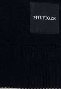 Maglione lavorato a maglia navy con orlo a coste testurizzato, dotato di un'etichetta nera rettangolare che riporta la scritta "HILFIGER" in lettere maiuscole bianche.