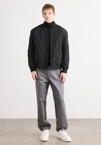 Μαύρο καπιτονέ μπομπέ μπουφάν με ριγέ μανσέτες, συνδυασμένο με γκρίζες παντελόνες και λευκά αθλητικά παπούτσια. Απλός, μοντέρνος σχεδιασμός με υφή.