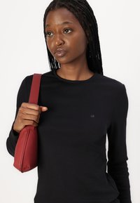 Chemise noire à manches longues avec un col rond et un petit logo gris. Le mannequin tient un sac à main rouge compact au design structuré.