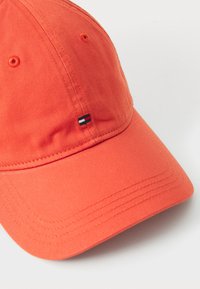 Oranžinė medvilninė beisbolo kepurė su išlenktu kraštu, ant priekinės dalies yra mažas juodas, baltas ir raudonas logotipo pleistras. Lygi tekstūra.