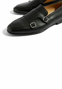 Crne kožne mokasine elegantnog dizajna, s dvostrukim srebrnim kopčama i dugim vrhom. Teksturalna završna obrada i minimalno šivanje.
