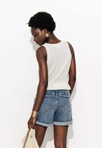 Débardeur en tricot blanc avec un motif texturé, associé à un short en denim roulé. Les accessoires comprennent de grandes boucles d'oreilles en or et un bracelet.