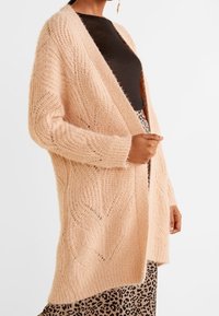 Vaaleanpunainen neulottu neuletakki, jossa on tilava mitoitus, ja jossa on teksturoitu lehtikuvio sekä avoin etuosa. Yhdistetty mustan paidan ja leopardikuosiset hameen kanssa.