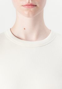 Wit geribbeld stoffen top met een ronde halslijn. Close-up dat de gladde textuur en subtiele stiksels toont. Kleine moedervlek op de nek.