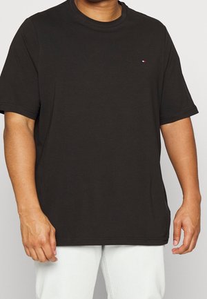 Person iført en ensfarvet sort T-shirt med korte ærmer og et lille rødt, hvidt og blåt logo på venstre bryst, kombineret med hvide bukser.