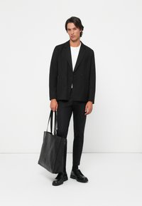 Blazer negro de corte cruzado con solapas en pico, combinado con una camiseta blanca y pantalones negros a medida. Hombre sostiene un bolso de mano de cuero negro.