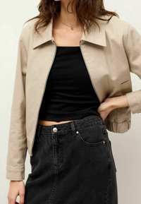 Veste beige courte avec un col, fermeture éclair à l'avant et poignets élastiques, portée sur un top noir et associée à une jupe en denim noire taille haute.