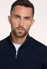 Maglione a zip di colore blu navy realizzato in maglia testurizzata, con colletto rialzato e dettaglio della cerniera in argento. Indossato sopra una camicia bianca.