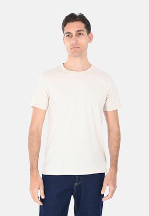 Giovane uomo che indossa una maglietta beige chiaro tinta unita a maniche corte e jeans blu scuro in piedi davanti a uno sfondo bianco.