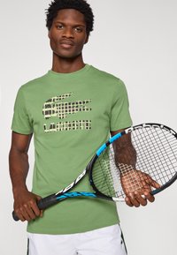 Πράσινο βαμβακερό μπλουζάκι με το λογότυπο Lacoste σε σχέδιο παραλλαγής. Το μοντέλο κρατά ρακέτα τένις με μαύρη και μπλε λαβή. Μινιμαλιστικός σχεδιασμός.