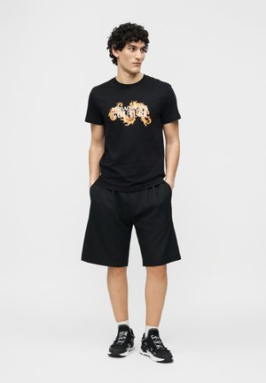 Jauns vīrietis ar cirtainiem matiem, valkājot melnu grafisko T-kreklu, melnas šortus un melnas sporta kurpes, stāv ar rokām kabatās vienkāršā fonā.