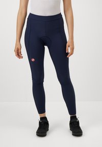 Sieviete, tērpta tumšzilgos polsterētos riteņbraukšanas legingos un melnās sporta kurpēs, stāv uz baltas fona.