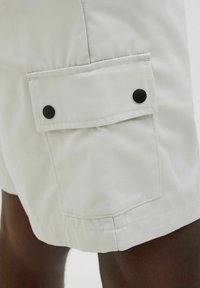Shorts beige clair en tissu lisse, avec une poche latérale à rabat et deux boutons noirs, dotés d'ourlets cousus pour plus de durabilité.
