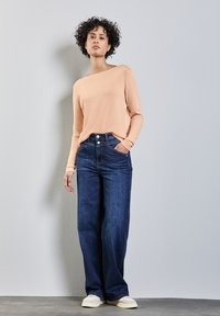 Helles Pfirsich-farbenes Langarmoberteil, kombiniert mit hoch taillierten, weiten dunklen Jeans. Zu den auffälligen Merkmalen gehören zwei Knöpfe und seitliche Taschen.