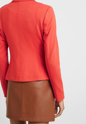 Kobieta w dopasowanej czerwonej marynarce i brązowej skórzanej spódnicy, widoczna od tyłu na jednolitym jasnym tle.