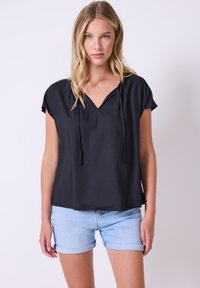 Donna con lunghi capelli biondi che indossa una maglietta nera a maniche corte e larghe e pantaloni shorts di denim blu chiaro arrotolati, in piedi su uno sfondo semplice.