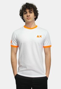 T-shirt bianco in cotone con dettagli arancioni sul colletto e sulle maniche, caratterizzato da un piccolo logo sul petto e un numero su una manica.