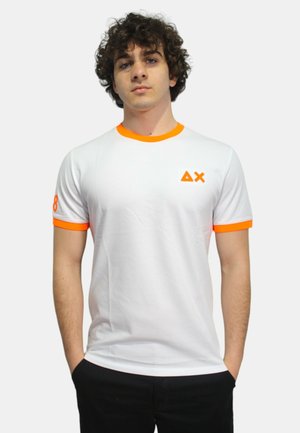 T-shirt bianco in cotone con dettagli arancioni sul colletto e sulle maniche, caratterizzato da un piccolo logo sul petto e un numero su una manica.