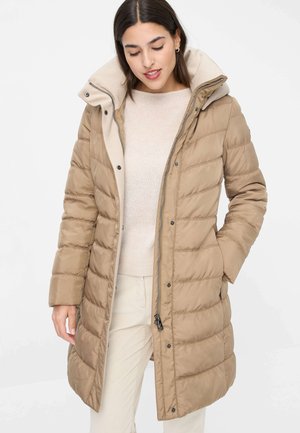 Frau trägt eine beige gesteppte lange Daunenjacke mit hohem Kragen, die mit Reißverschluss und Knöpfen geschlossen ist, kombiniert mit einem hellen Pullover und cremefarbenen Hosen.
