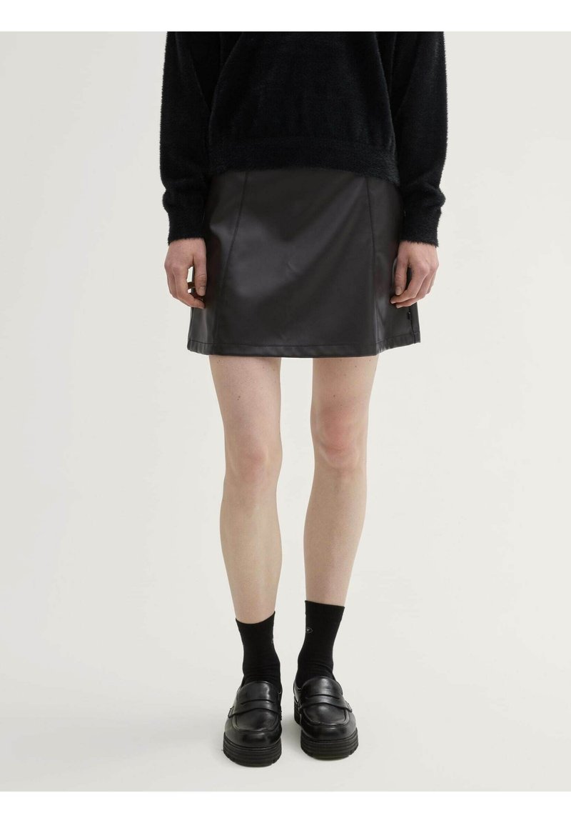 Minifalda de cuero negra con una textura suave, que presenta un corte sencillo y detalles de costura visibles. La lleva puesta con un suéter negro y mocasines.