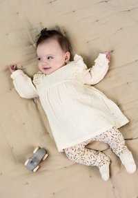 Robe couleur crème avec des détails en volants et un tissu texturé, associée à des leggings à motifs avec des designs floraux ; une petite voiture en jouet grise à proximité.