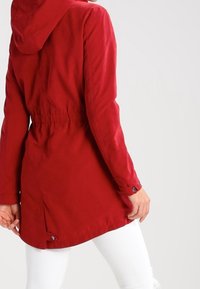 Röd huva jacka med cinch midja, långa ärmar och justerbara manschetter; har en rak passform och delad fåll bak.