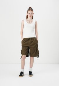 Hvit ribbet tanktopp, olivengrønne cargo shorts med sidelommer og snor, paret med svarte joggesko og hvite ankelsokker.