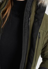 Olivgrön isolerad jacka med avtagbar huva i päls, svart foder och dragkedjedetaljer. Texturerad tyg med sydda detaljer.