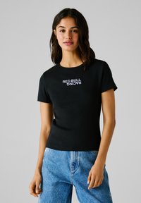 Schwarzes, geripptes T-Shirt mit kurzenÄrmeln, das das weiße, gestickte Logo "RED BULL RACING" zeigt. Kombiniert mit hellblauen Jeans.