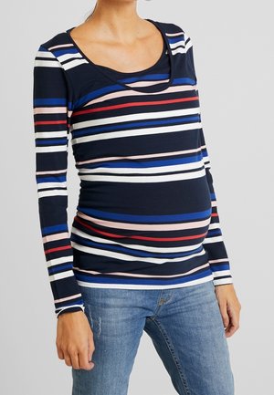 Zwangere vrouw die een marineblauwe lange mouw top draagt met rode, witte, roze en blauwe horizontale strepen en blauwe spijkerbroek.